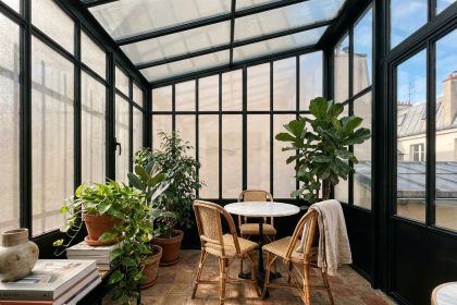 Verrière industrielle noire intégrée dans un appartement parisien avec sol en terre cuite, table en marbre et plantes vertes luxuriantes sous une structure en acier et verre
